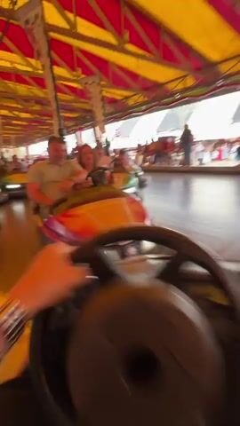 The video shows a first-person point of view of a person driving a bumper car. The hands of the driver are visible on the steering wheel. The ride is full of other bumper cars with various drivers and passengers. The ceiling is striped with red and yellow colors. The ride appears to be indoors.