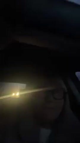 The video shows a woman wearing glasses and a gray coat sitting in a car. She is singing and mouthing the words to a song.  A streetlight is visible in the background. The sky is dark.