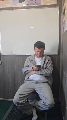 The video appears to be an informal recording, likely capturing moments in an office or communal space in Jararyk, Uzbekistan. The setting is utilitarian, with a whiteboard on the wall, filing cabinets, shelves filled with documents and binders, and a desk with a computer and printer. The lighting suggests it could be daytime, with natural light coming from a window.

There are at least three men visible. The first, at the beginning of the video, is sitting in a chair, dressed in a light grey jacket and pants, looking at his phone. The second man, in the middle portion, is seen from behind, wearing a white t-shirt and grey pants, sitting at a desk and looking towards a computer screen. The third man, towards the end, is wearing a white t-shirt and a black baseball cap. He looks directly at the camera and gives a thumbs-up, with a slight smile.

Objects of note include a white teapot with decorative patterns and small cups on a desk, a large water cooler bottle, and various papers and documents on the walls and shelves. Some of the papers appear to be charts, diagrams, or instructional materials, but the text is not clearly legible. The overall atmosphere seems relaxed, possibly a break in work or a casual check-in.