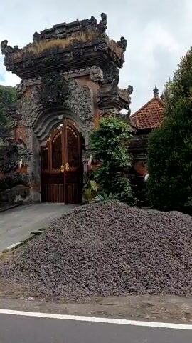 The video shows a roadside view of a village. In the foreground, a paved road runs horizontally across the bottom of the frame. To the left of the road, a pile of logs is visible. Beyond the logs, a grassy area leads to a low stone wall. Behind the wall, a building with traditional Balinese architecture is seen, featuring intricate stone carvings and a tiered roof. A lush green tree stands in front of the building. Further to the right, another traditional entrance gate is visible with a person walking up the stairs. Behind this gate, a multi-story building with balconies can be seen. Further down the road, a covered structure houses several stone sculptures and architectural elements, likely for sale or display. The sky above is partly cloudy.