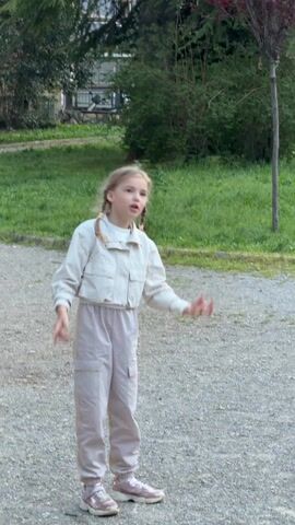 A young girl with blonde hair in pigtails, wearing a white jacket, beige pants, and pink sneakers, stands on a gravelly surface in an outdoor setting. Behind her is a patch of green grass and some trees. The girl dramatically exclaims, "Тут летучие мыши!", which translates to "There are bats here!", and then she screams, clasping her hands to her chest.