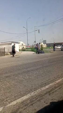 In a daytime outdoor scene, a group of four individuals are engaged in an interaction near a crosswalk. One individual, a man wearing a dark shirt and carrying a black bag, walks from left to right across the frame, away from the group. In the center of the frame, three individuals are gathered around what appears to be a person lying on the ground, possibly being assisted or restrained. One of the individuals appears to be wearing dark clothing, another a light-colored shirt, and the third a darker shirt. A silver hatchback car is visible driving past in the background from left to right. The environment suggests an urban or suburban setting with a road, sidewalk, traffic signals, and buildings in the distance. The lighting indicates it is daytime, with a clear sky.