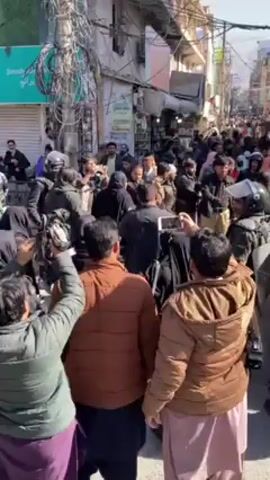 A crowded street scene shows many people packed into a narrow street, lined on both sides with multistory buildings. The scene is shot at eye-level from behind several people, at least one of whom is recording the event on a cell phone. Several members of the crowd are wearing riot gear, including helmets and shields, while most others are in civilian clothing, including coats and jackets. Some bystanders are observing and recording the event, while others are pushing forward or engaging with those in riot gear. A tangle of wires hangs above the street.