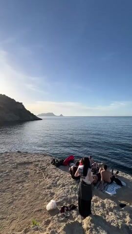 A woman with dark hair, wearing a white t-shirt and dark skirt, stands on a light-colored rocky outcrop, holding a phone and facing a group of people. Below her, on a lower rock platform at the water's edge, approximately four to five individuals are seated, some on towels, relaxing. The scene is a coastal landscape with a clear blue sea under a bright blue sky, featuring distant landmasses on the horizon to the left. The camera pans slowly from left to right and down, revealing the rocky cliff face, the calm blue water with small waves breaking against the shore, and the curved coastline. Bright sunlight illuminates the scene.