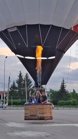 A hot air balloon is being prepared for flight outdoors. The balloon is white with colorful geometric patterns near the top. It is tethered and the burner is actively firing, sending a large flame into the balloon. A wicker basket is suspended beneath the balloon, and four people are visible inside. Two people are in the front of the basket, with one waving. The other two people are in the back of the basket. The balloon is on what appears to be a paved surface, possibly a parking lot or street, with white markings on the ground. There are cars and trees in the background, and the sky is visible, with clouds and hints of sunset colors.