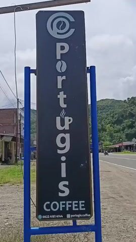 The video shows a vertical sign for "Portugis Coffee" located in Gleputoh, Indonesia. The sign is black with white lettering and features a stylized coffee bean and a steaming coffee cup. The name "Portugis" is written vertically, with each letter appearing on a new line, and the word "COFFEE" is written horizontally below it. The sign is supported by blue metal poles and stands beside a paved road. In the background, there are buildings, some with open storefronts, and lush green hills under a cloudy sky. A few motorcycles are visible on the road, suggesting it's daytime. The overall scene depicts a roadside coffee shop in a semi-rural or suburban area. The visible text on the sign includes "Portugis COFFEE," a phone number "0822 4161 4144," and a website address "portugis.coffee". The weather appears to be overcast.