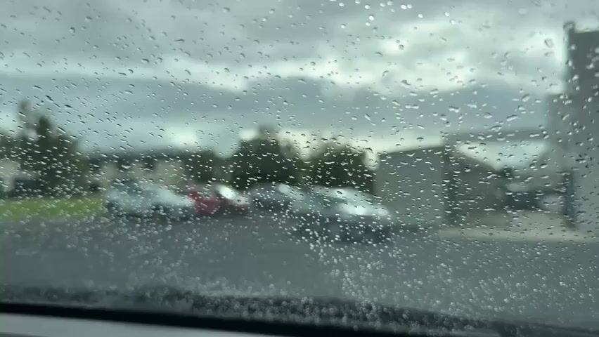 This is a close-up shot of a car windshield covered in rain droplets. The background is blurred and shows a parking lot with several cars parked. The sky is overcast and cloudy, indicating rainy weather. The overall scene depicts a gloomy and wet atmosphere.