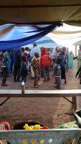 Several children and adults mill about in a dusty, open area, partially shaded by a tent-like structure with a draped blue sheet. A wooden table with a water bottle and some food on it stands in the foreground. Most of the people are wearing colorful clothing, and some appear to be dancing or moving to music. The overall atmosphere seems celebratory or festive.