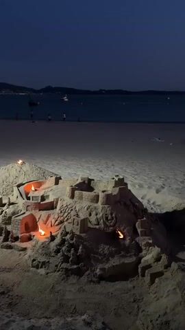 The video shows a sandcastle illuminated by candles on a beach at night. The sandcastle is elaborate, with multiple towers, walls, and archways. Some of the candles are placed inside the sandcastle, casting a warm glow on the sand structures. In the background, the ocean is visible, with distant lights from a coastline or boats. The sky is dark, indicating it is nighttime. There are a few silhouetted figures walking on the beach in the distance. The camera is positioned at a low angle, focusing on the sandcastle.