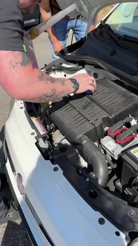 A person with tattoos on their left arm and wearing a black t-shirt and grey pants is seen pouring a pink liquid from a yellow bottle with a white hose into the engine compartment of a white car. Their right hand is holding the bottle and hose while their left hand is on the engine. Another person in the background is wearing a brown shirt and blue jeans. The car's hood is open, revealing the engine. The environment appears to be outdoors, with sunlight visible.