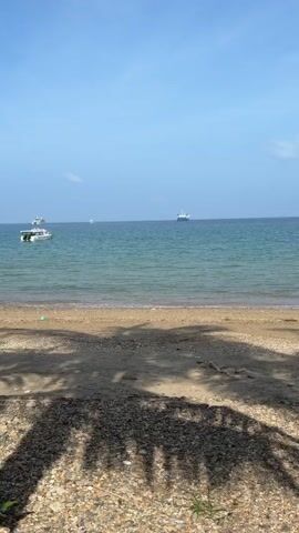 The video shows a view of a calm ocean from a sandy beach. The water is a greenish-blue, and there are small waves lapping at the shore. Several boats are visible on the horizon, including a white speedboat on the left and a larger white ship further out. The beach in the foreground is covered in sand and small pebbles, with palm tree shadows cast across it. The sky is a clear, bright blue, indicating a sunny day. There are no people visible in the video. The camera appears to be stationary, capturing a static scene.