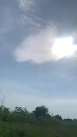 The video shows a sunny sky with white clouds partially covering the sun. A line of trees and bushes is visible along the bottom of the frame. Someone is talking in the background, expressing surprise at how bright the sun is.