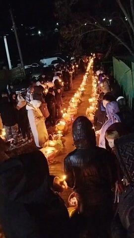 The video shows a nighttime scene of people gathered outdoors, holding lit candles and baskets. The crowd forms a long line, creating a pathway illuminated by the candlelight. People of varying ages and attire are present, some wearing hooded jackets or white robes. The environment includes visible streetlights, parked cars in the background, a green fence, and a paved ground surface. The activity appears to be a religious procession or gathering, with a solemn and reverent atmosphere.