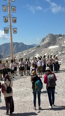The video captures a brass band playing in a mountainous landscape. The band members are dressed in traditional Bavarian attire, including lederhosen and hats. They are positioned in front of a tall wooden pole adorned with various shields featuring illustrations. Behind the band, snow-capped mountains rise against a clear blue sky. Several spectators, including people with backpacks, are observing the performance from a gravelly area in the foreground. The scene appears to be a cultural event or festival taking place at a high altitude.