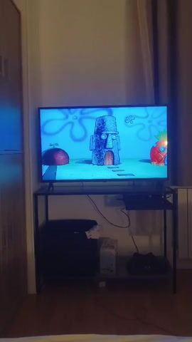 A television displays an animated scene from the show SpongeBob SquarePants. On the left is SpongeBob, and on the right is Squidward. They appear to be in a room with a red, textured wallpaper. SpongeBob is smiling and looking at Squidward, while Squidward has a surprised expression on his face and is holding a book that is open. There is a desk or table with a stool next to Squidward. The TV is placed on a black metal stand. To the left of the TV stand, there is a black object and a white computer tower. To the right of the TV stand, there is a dark object, possibly a shelf with items on it, and a white radiator. The room has wooden flooring and light-colored walls.
