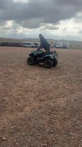 The video captures two individuals riding a green all-terrain vehicle (ATV) across a wide, gravelly expanse. The driver, clad in a dark jacket, is initially standing on the ATV before settling into a seated position. A passenger, also in a dark jacket, waves an arm while riding. In the background, several white and black vans are parked, suggesting a gathering point or parking lot for a tour group. Other people are visible near the vans. The setting is an open, dusty area with a distant, undulating landscape under a heavily clouded sky. The weather appears overcast, indicating daytime but not necessarily bright sunshine. The scene is consistent with a recreational activity, possibly a quad biking tour in Lalla Takarkoust, Morocco. There is no visible text.