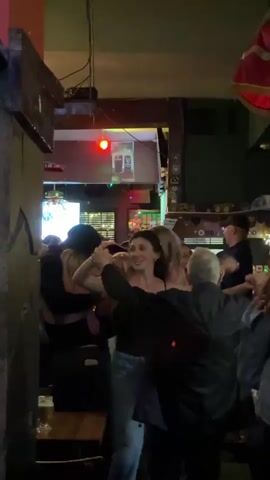 The video shows a group of people dancing and singing in a crowded bar. The lighting is dim, and there is a red light visible on the ceiling. The people are dressed casually. One woman in the center is wearing a black tank top and jeans, and she is singing loudly. The music appears to be upbeat and energetic. There is a beer glass on a wooden table in the foreground. There is a television screen in the background displaying video content. There is a Guinness sign on the wall. A flag hangs in the background.