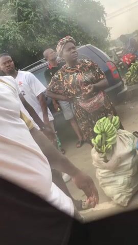 A group of people are gathered outdoors, possibly at a market. In the foreground, a person wearing a white shirt is partially visible, with their arm extended. To their right, a woman wearing a patterned dress and headwrap stands with her hands on her hips, looking to her right. She has a fanny pack around her waist. Next to her, a large sack of green bananas is on the ground. Behind her, a dark-colored vehicle is parked. Other individuals are visible in the background, some blurred. The environment appears to be somewhat dusty or hazy.