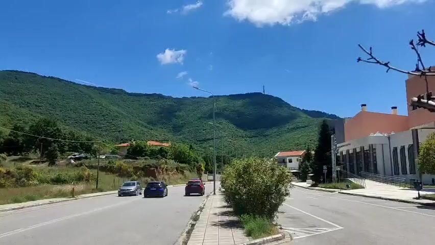 The video shows a view of a road in a town or village with a mountain in the background. The sky is bright blue with some scattered clouds. There are several cars driving on the road.  A building is visible on the right side of the road. There are trees and bushes along the side of the road. The mountain is covered with green vegetation. A utility pole with wires is visible on the left side of the road.