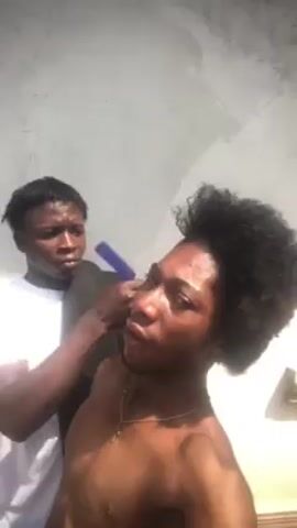 A shirtless brown-skinned man with short, curly black hair stands outside and is being combed by another brown-skinned man with short, straight hair wearing a white t-shirt. The first man looks at the camera as his hair is combed, occasionally tilting his head. The men stand in front of a white wall in an outdoor environment.
