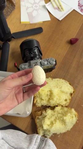 A hand holds a small, white, egg-shaped object with a textured surface. The object appears to be made of a solid, creamy substance. The hand is adorned with two silver rings. In the background, there is a section of yellow, fluffy bread, a camera, a small container holding additional similar white objects, and miscellaneous items scattered on a wooden surface. The person wearing the sweatshirt appears to be documenting the object with the camera on the table.