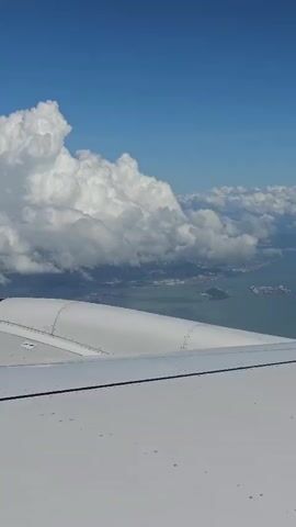 The video is an aerial view from an airplane window. The foreground is dominated by a portion of the airplane's wing, which is white and smooth with visible rivets along its edge. Beyond the wing, the scene opens up to a vast expanse of blue sky dotted with large, fluffy white clouds. Below the clouds, a body of water, likely the ocean, is visible, with a coastline and urban structures in the distance. Several ships or boats can be seen on the water near the coast, suggesting a port or harbor. The lighting indicates it is daytime.