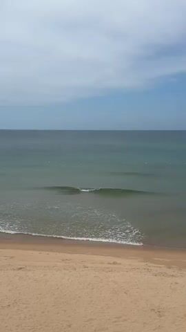 A gentle wave crests and breaks on a sandy beach. The ocean stretches out to the horizon under a partly cloudy sky. The water is a calm, greenish-blue, and the sand is light brown. The scene is serene and tranquil.