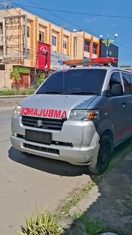 The video shows a silver Suzuki ambulance parked on the side of a street. The word "AMBULANCE" is written in red letters on the hood of the vehicle, and it has a red and orange light bar on the roof. A motorcycle passes by in the background. The background includes buildings and a clear blue sky. The ambulance is parked on asphalt next to a curb with some grass.