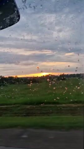 A video clip shows a view through a window, possibly a car, with water droplets on the glass. The scene outside is a rural landscape with green fields and trees under a cloudy sky during what appears to be sunset. The sun is visible on the horizon, casting a warm glow. There are some man-made structures in the distance, including what looks like a fence and some abstract sculptures or playground equipment. The camera is moving, suggesting it is filmed from a vehicle. The overall impression is one of a peaceful, scenic drive.