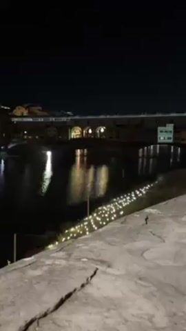 At night, the Ponte Vecchio bridge in Florence, Italy, is illuminated with a light show. The bridge’s buildings reflect in the Arno River. Along the near shoreline, decorative lights are visible. The camera is situated on a stone wall, displaying its rough texture. No people are readily visible.