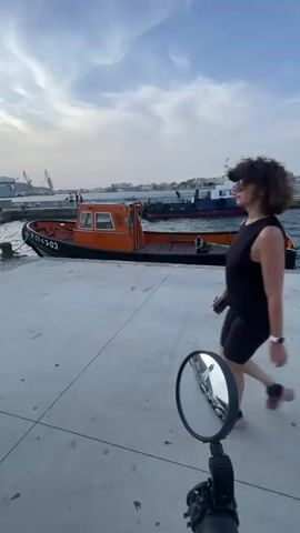 A person is walking away from the camera on a concrete pier. They are wearing a black tank top, black shorts, and pink and white sneakers. A round mirror is held in the foreground, reflecting the scene. In the background, an orange boat is docked, and behind it, a blue and white tugboat is at anchor. The water is calm, and the sky is overcast. Buildings and cranes are visible in the distance on the shoreline.