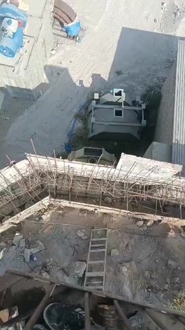 An aerial view captures a construction site with various structures and materials. In the foreground, a partially constructed building features scaffolding and exposed rebar. A wooden ladder is propped against the foundation, suggesting ongoing work. Beyond this, a larger, grey industrial-looking structure stands, surrounded by piles of dirt and debris. Further in the background, blue tarp-covered objects and more piles of material are visible. The overall impression is a busy, active construction environment.