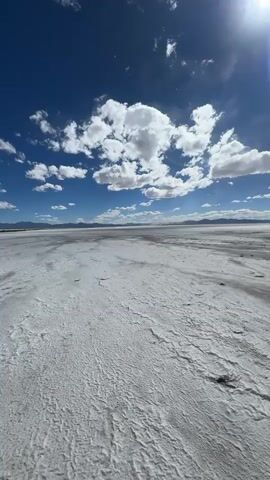 This video showcases a vast, sun-drenched salt flat under a brilliant blue sky dotted with fluffy white clouds. The ground is a stark white expanse, textured with ripples and patches of dried mud, stretching out to a distant horizon. In the far distance, a low mountain range is visible, hinting at the arid landscape of Tres Morros, Argentina.

The bright sunlight suggests it's midday, and the clear sky with scattered clouds indicates fair weather. There are no people or man-made objects visible, emphasizing the natural, desolate beauty of the location. The overall impression is one of immense space and quietude, a serene and almost otherworldly environment. The camera is positioned low to the ground, giving a ground-level perspective of the expansive salt flat, with the sky filling the upper portion of the frame. The lack of any discernible activity or human presence contributes to a feeling of solitude and the raw power of nature. No text is visible in the media file.