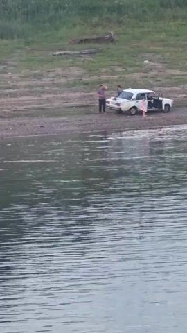 In this video, a group of people are gathered near a white car parked on a muddy, gravelly shore next to a large body of water. The scene appears to be a natural landscape with a grassy embankment in the background. The time of day seems to be late afternoon or early evening, given the soft lighting. The weather appears calm, with gentle ripples on the water's surface.

There are at least three people visible near the car. One person, a man in a dark shirt and pants, stands to the left of the car, facing away from the camera. Another person, a child in a light-colored dress, is near the open driver's side door. A third person, a child wearing a light-colored garment, is walking towards the water's edge. Further down the shore, another individual is standing in the shallow water. The overall atmosphere suggests a casual outing or a break during a journey. There is no visible text in the video.