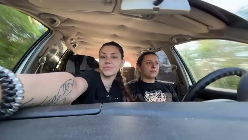 The video shows two young women in the front seats of a moving car. The woman in the passenger seat, wearing a black shirt, looks at the camera and makes a kissy face. She has a tattoo on her left arm and a beaded bracelet. She later puts her hair in a ponytail. The woman driving, also wearing a black shirt, keeps her eyes on the road.  The car's interior is visible, including the dashboard, steering wheel, and a cluttered back seat. Trees pass by outside the car windows, suggesting the car is moving through a wooded area.  Music is playing in the background.