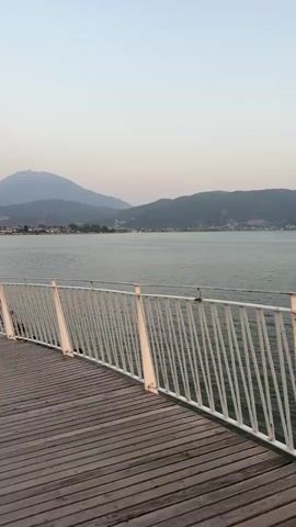 The video shows a wooden pier with a white railing along the side. The pier extends over a body of water, with a town and mountains in the background. The sky is a pale, hazy color, suggesting either early morning or late afternoon. Several people are visible walking along the pier and on the distant shoreline. One woman in a black dress is walking away from the camera on the left side of the pier. The water appears calm, and there are some palm trees on the left side of the pier. The overall atmosphere is peaceful and serene.