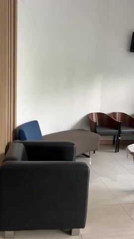 The video pans across a waiting room with tiled floors and white walls. In the foreground, there are two black leather couches. To the left, partially visible, is a wooden door frame. In the middle of the room, there is a long brown bench with a blue cushion on one end. Further back, against the wall, are three curved wooden chairs with black seats. A small wooden coffee table with a white lace doily and two remote controls sits in front of the chairs. A small black television is mounted on the wall above the chairs, with visible wires. To the right, a large window with multiple panes lets in natural light, showing greenery outside. A white roller blind is pulled down over the top portion of the window.