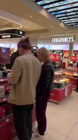 The video shows a busy duty-free shop in an airport. Signs for brands like Chanel, Tom Ford, and Dior are visible. People are browsing and shopping at the various product displays, which contain fragrances, cosmetics, and other beauty items. A man with a bun stands near a Tom Ford display looking at items. In another shot, a man in a suit and a woman in a blue shirt are talking near a Chanel display.