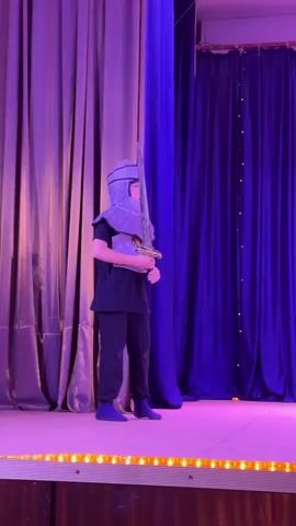 The video shows a young person dressed in a knight's costume on a stage. The costume includes a helmet, a chest plate, and a sword. The person is standing on a stage with purple curtains in the background. The stage is lit with purple and pink lights. The person is holding the sword and appears to be performing or practicing. There is a white box or prop on the right side of the stage. The person moves the sword in a circular motion and then holds it in front of them.