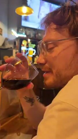 The video shows a man drinking a glass of red wine in what appears to be a pub or bar setting. He has reddish hair, a beard, and wears glasses. There's a tattoo on his left forearm. The camera pans to another man, bald with a beard and glasses, who is gesturing and talking. A shot glass is visible on the table. The background includes bar decorations, a television screen, and lighting fixtures.