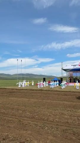 A group of people in traditional white and red outfits are performing a dance in front of a stage in a field. A person on horseback wearing a green shirt and hat is facing away from the camera, holding a long pole. The sky is blue with white clouds.