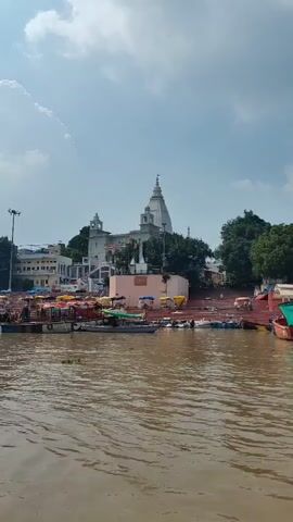 A vertical shot captures a wide, muddy river in the foreground. Several small boats are moored along the bank on the left side. Beyond the riverbank, a white temple with a spire dominates the scene. The temple is surrounded by trees and other buildings. The sky above is partly cloudy. The camera moves slowly to the right, revealing more of the boats and the temple complex.