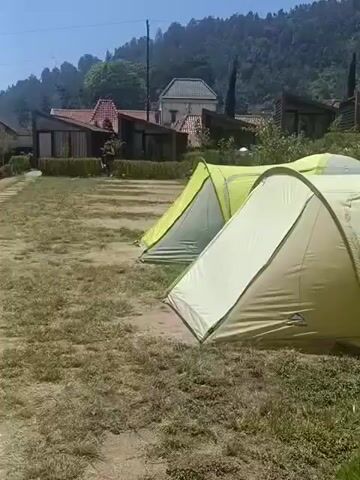 The video shows a campsite with several tents set up on a grassy area. In the foreground, there are two large tents, one predominantly light green with a tan accent and the other a lighter shade of green. A red tent is visible behind them, and another white tent is further in the background. The campsite appears to be situated in a hilly or mountainous area, with trees and buildings visible in the distance. The sky is clear and blue, suggesting a sunny day. There are no people visible interacting with the tents or the environment in this short clip. The camera pans slightly, showing different angles of the campsite.