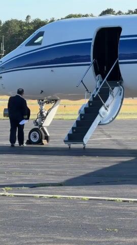 A pilot in a dark uniform is conducting a pre-flight inspection of the landing gear of a private jet. The jet is white with a blue stripe. The cabin door is open, and a set of stairs is extended. The pilot is bending down to examine the front landing gear, holding a document in his left hand. The aircraft is parked on an asphalt tarmac, with a grassy area and trees visible in the background. The sky is clear and blue, indicating daytime.