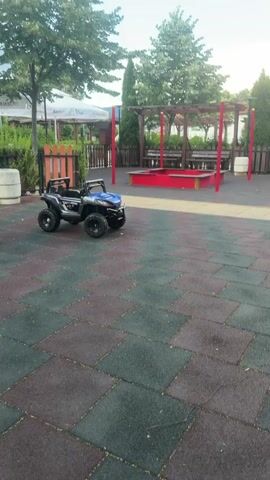 This is a vertical video of an empty playground. The ground is covered in interlocking rubber tiles in shades of green, black, and red. In the foreground, to the left, is a yellow plastic slide. To the right of the slide, in the center of the frame, is a small, black and blue electric ride-on toy car, resembling an ATV. In the background, there is a red sandbox with wooden borders and benches. Behind the sandbox is a wooden pergola. Further in the background, there are trees, a fence, and buildings with white canopies. The sky is blue with some clouds. The overall impression is that of a public outdoor play area.
