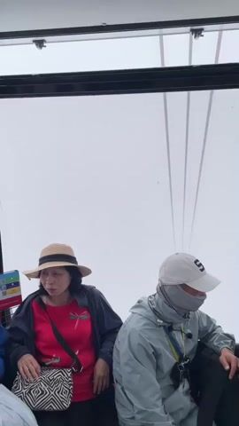 The video shows the interior of a cable car with several passengers. A woman wearing a straw hat and a red shirt is visible on the left. Next to her, a person in a light blue jacket and a white baseball cap with a face covering is seated. Further to the right, another person in a dark jacket is seated, facing away from the camera. The cable car's windows offer a view of the sky, which appears overcast and foggy, with the thick cables of the cable car system visible against the white background. The overall environment suggests a journey in a mountain or elevated area.