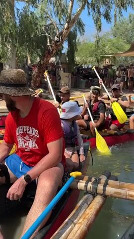 The video shows a group of people in canoes on a body of water, likely a river. Several canoes are visible, with people paddling. In the foreground, a man wearing a red t-shirt and a straw hat is in a canoe. He has a beard and is wearing blue shorts and a watch. A child in a blue hat and life vest is sitting in front of him. Behind them, other canoes are filled with people, many wearing life vests and holding paddles. The individuals in the canoes appear to be actively paddling. The background features lush green trees and vegetation, with a clear blue sky overhead. There are also some structures or buildings visible in the distance, possibly part of a dock or recreational area, with other people gathered there. The water appears somewhat choppy around the canoes, indicating movement. The overall scene depicts an outdoor recreational activity, likely a group canoeing trip.