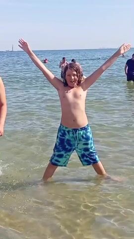 A young boy with long, light brown hair stands in shallow ocean water with his arms outstretched to his sides, forming an X shape. He is wearing blue and green tie-dye swim trunks. Waves lap around his legs, creating splashes. In the background, several other people are in the water, some swimming and others standing. The sky is clear and blue, indicating a sunny day. The water is clear and appears shallow.