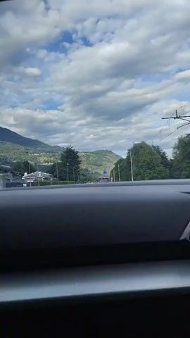 The video shows a view from inside a vehicle, looking out at a road with mountains and trees in the background. The sky is cloudy. There are several cars on the road, suggesting a moderate amount of traffic. The scene appears to be set in a rural or semi-rural area with houses and streetlights visible on the left side of the road. A triangular road sign with a red border and a white background, depicting a traffic hazard, is visible on the right side of the road. The overall impression is of a daytime drive through a scenic landscape. There is no visible text in the video.