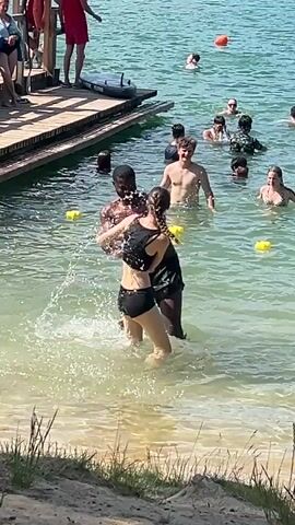 The media file shows a group of people at a body of water, likely a lake or beach, on a sunny day. The water is clear and shallow near the shore, with a sandy bottom visible. In the foreground, two individuals, a man and a woman, are interacting playfully in the water. The woman is wearing a black bikini top and bottoms, while the man is wearing black swim trunks. They appear to be engaged in some sort of playful game or dance, with water splashing around them.

In the background, several other people are visible in the water, some swimming and others standing. A wooden pier extends into the water on the left side of the frame, with more people gathered on it. There are also several yellow buoys floating in the water. The overall scene suggests a recreational setting, possibly a popular swimming spot during warm weather. The sunlight indicates it's daytime, and the clear skies suggest good weather. The presence of a pier and a sandy shore points towards a natural body of water rather than a swimming pool. No text is visible in the media file.